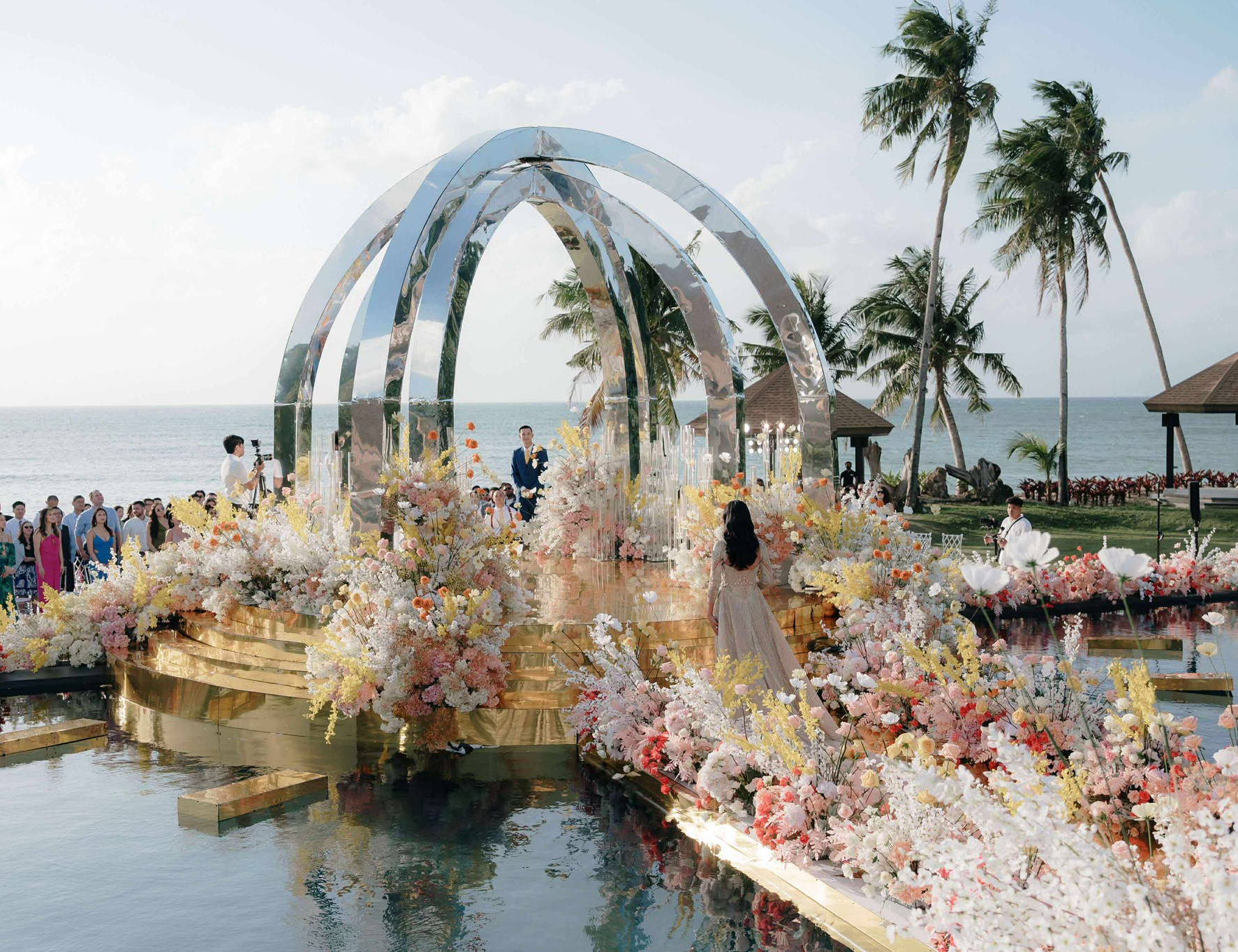 Bride walking down a gold aisle surrounded by lush pastel flowers, approaching a modern metallic wedding arch by the sea on Balesin Island.