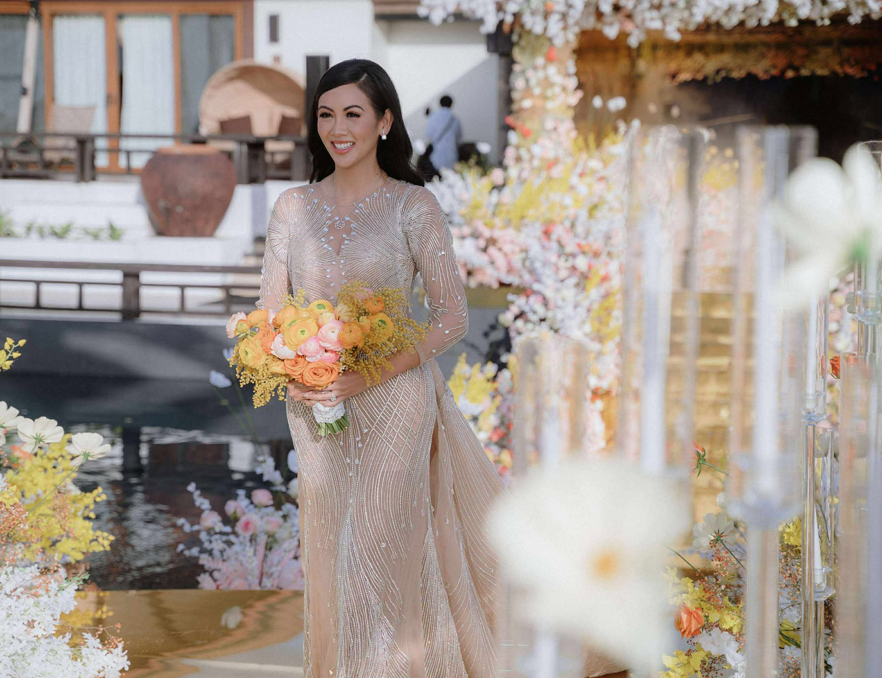 Bride in a sparkling gown holding a yellow and peach bouquet, smiling as she walks through the floral wedding aisle at Balesin Island.