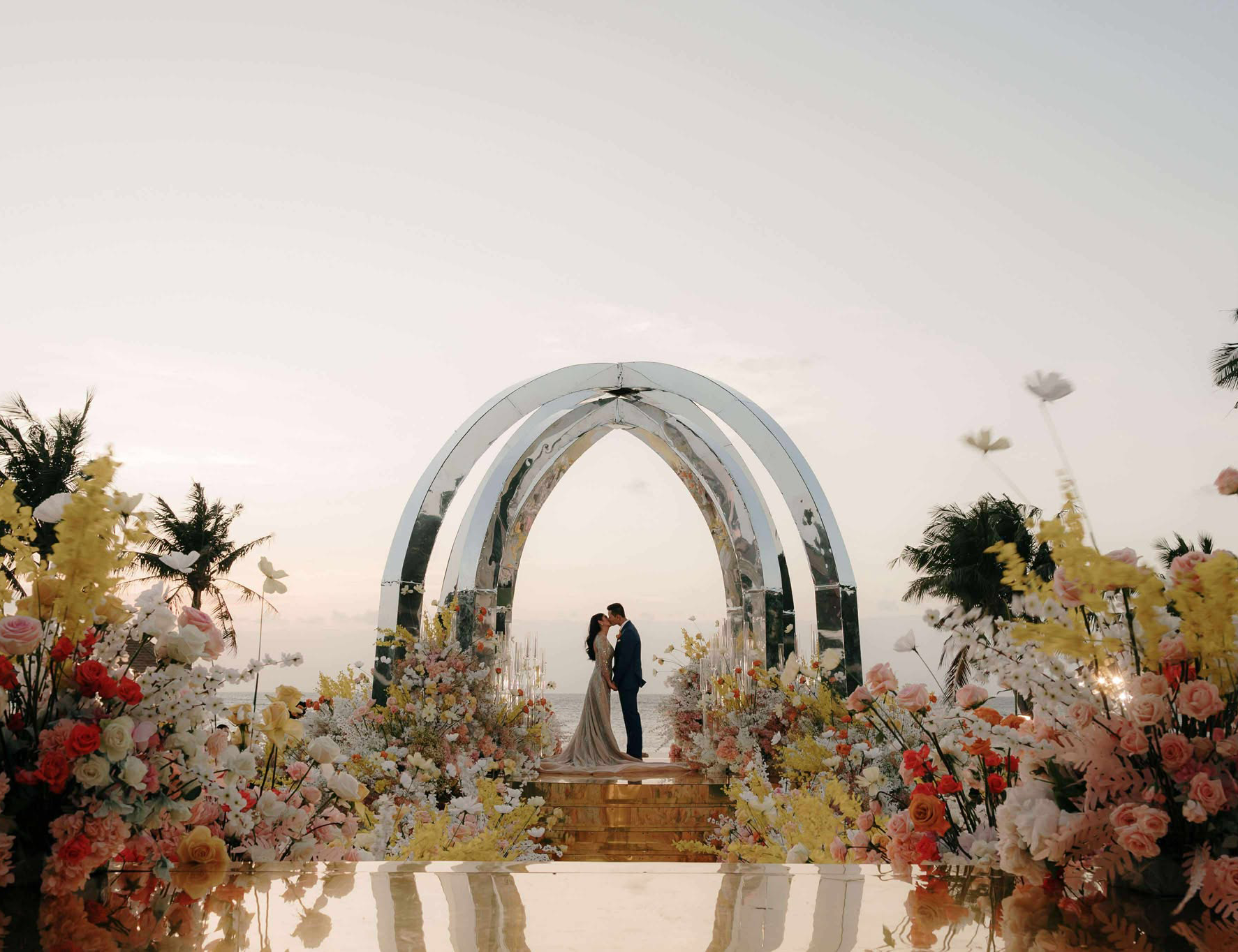 Bride and groom sharing a kiss under a modern silver arch, surrounded by pastel flowers and palm trees at sunset during their Balesin Island wedding.