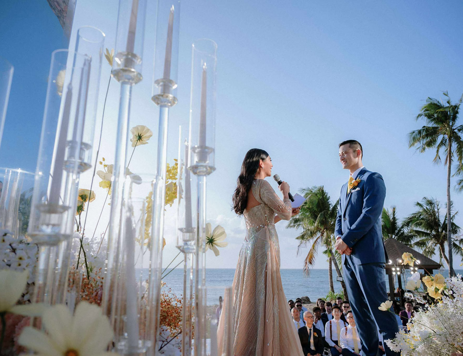 Bride and groom exchanging vows on a golden stage with tall glass candles and abundant pastel flowers, set against the sea and palm trees at Balesin Island.