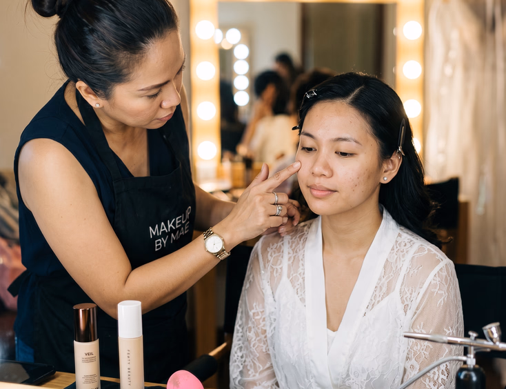 Close-up of Filipino HMUA testing a Filipino bride's skin texture before makeup application with primer, foundation, and airbrush tools arranged on the vanity beside a lighted mirror