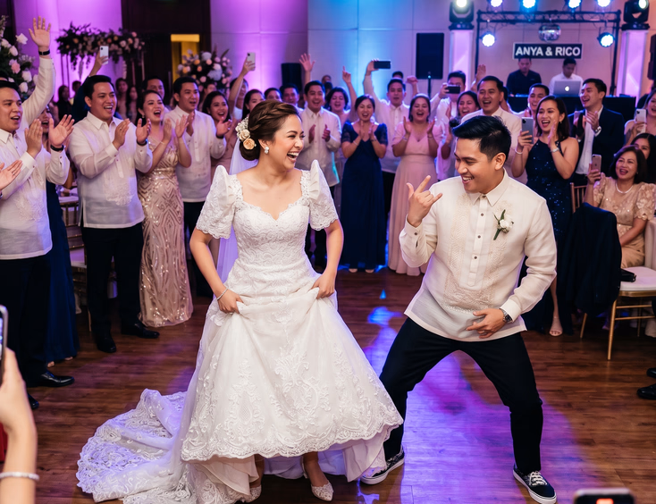 Filipino bride laughing while hiking up her gown and groom striking a playful hip-hop stance mid-transition from a slow first dance to an upbeat routine, with guests standing and cheering under purple and blue uplights