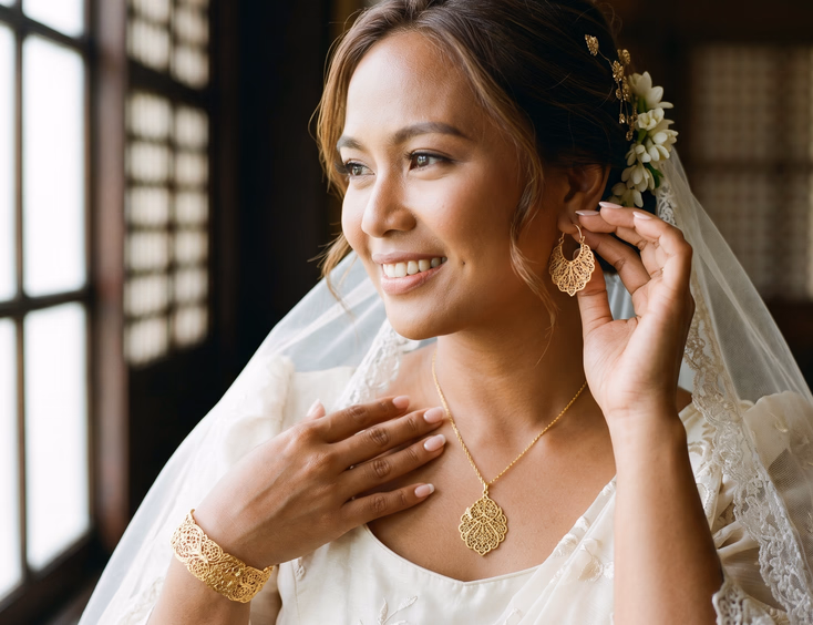 Close-up of Filipino bride wearing matching gold filigree kalado-pattern creolla earring, pendant necklace, and bangle, soft window light illuminating coordinated metalwork details across hands and jewelry, editorial wedding photography