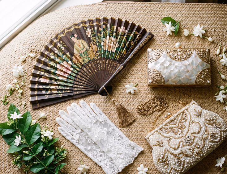 Overhead flat lay of Filipino bridal accessories on abaca mat including hand-painted abanico, white lace gloves, capiz shell clutch, and beaded ivory evening bag surrounded by sampaguita flowers, artisan product photography