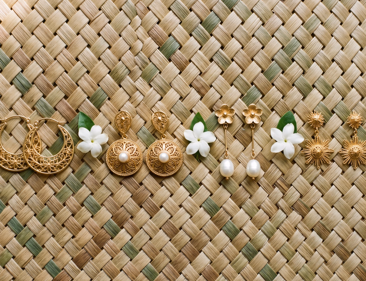 Flat lay of four pairs of Filipino heritage earrings on handwoven banig mat including gold filigree creolla hoops, tambourine earrings, pearl drop earrings, and sunburst studs with fresh sampaguita blossom, editorial jewelry photography