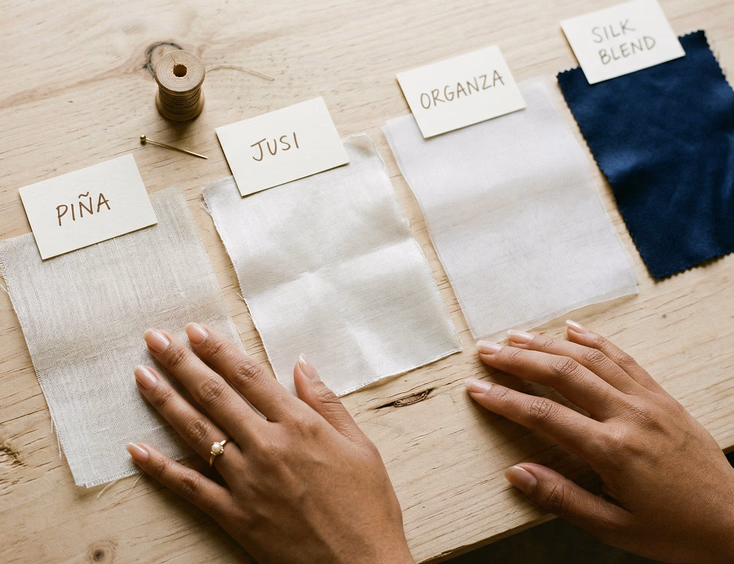 Flat-lay of four fabric swatches including piña jusi organza and silk blend on a light wooden surface with handwritten labels and Filipino woman's hands touching the piña fabric