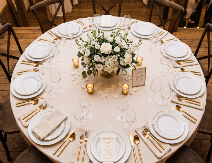 Overhead flat lay of a round Filipino wedding reception table with ten place settings, white plates, gold cutlery, cream napkins, white rose centerpiece, and printed menu cards