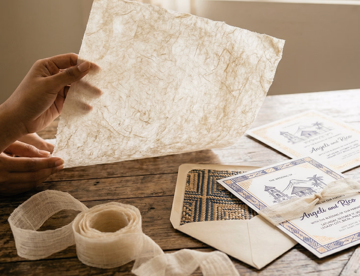 A close-up of a young Filipino woman's hands holding a sheet of natural abaca paper up to soft window light, with a piña cloth ribbon and banig-patterned envelope liner resting on the wooden surface below and a finished letterpress wedding invitation card visible in the background.