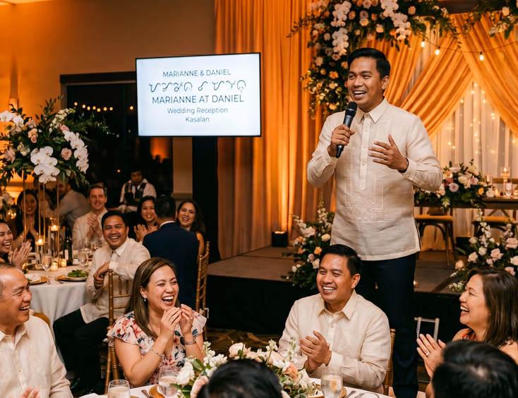 Filipino wedding host speaking animatedly on indoor reception stage before laughing mixed Filipino and foreign guests with couple's names on LED screen under warm amber lighting and floral draping