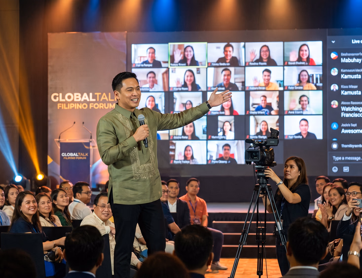 Filipino emcee on stage during a hybrid event gesturing to a screen displaying live chat and remote attendees, filmed by a camera crew.