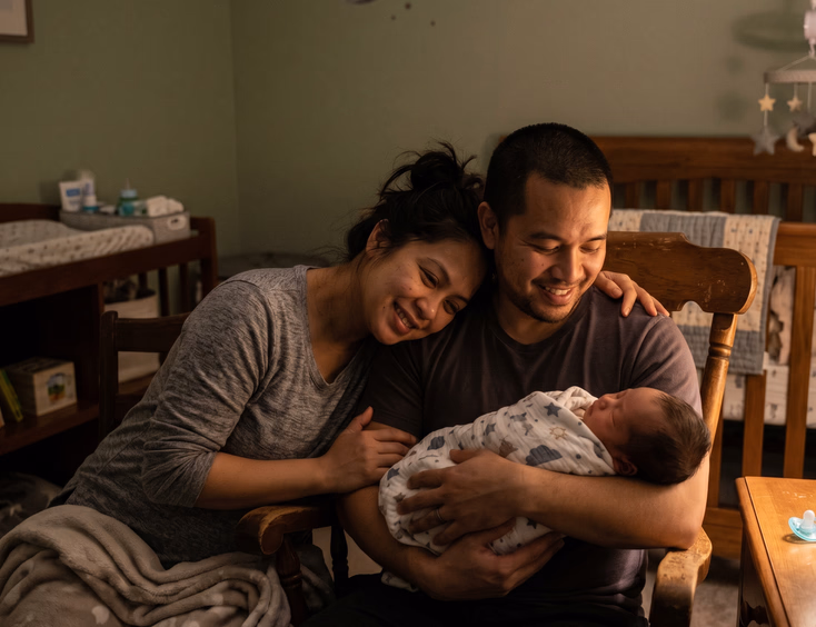 A tired but smiling Filipino couple shares a tender, exhausted teamwork moment in a dim nursery at night with their newborn under soft lamp light.
