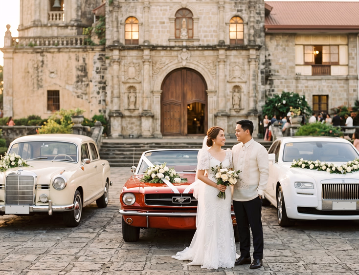 Three bridal cars parked in front of a Spanish colonial church including a cream vintage Mercedes-Benz, red Ford Mustang convertible, and white Rolls-Royce Ghost with a Filipino bride in white lace gown and groom in Barong Tagalog at golden hour