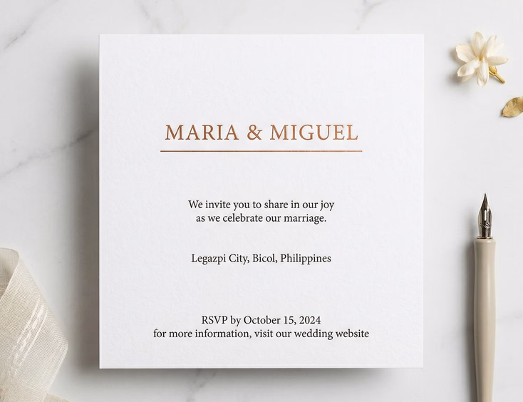 A close-up flat lay of a minimalist Filipino wedding invitation on thick white cotton cardstock featuring clean serif typography and a single copper foil name line, resting on a smooth white marble surface beside a single dried white bloom and a slim calligraphy pen in soft diffused natural light.