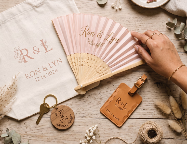 Flat lay of personalized Filipino wedding accessories on a light wooden surface including a monogrammed canvas tote bag, bamboo satin fan, leather bag tag, and engraved wooden keychain with a Filipino bride's hand reaching for the fan in soft natural daylight