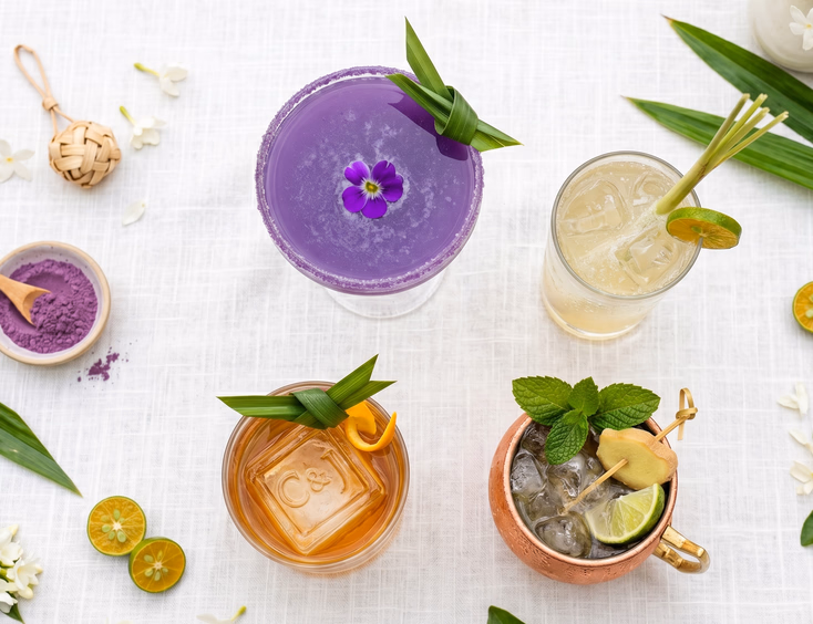 Top-down flat lay of four Filipino-inspired wedding cocktails including an ube martini, calamansi fizz, pandan old fashioned, and ginger mule with fresh local ingredients on a white linen tablecloth