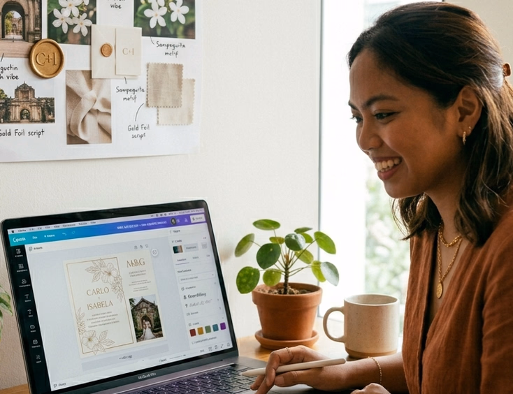 A Filipino woman in her late 20s sits at a bright desk with a laptop open to a wedding invitation design platform showing a partially customized digital invitation, with a printed reference mood board pinned to the wall behind her in warm natural light from a side window.