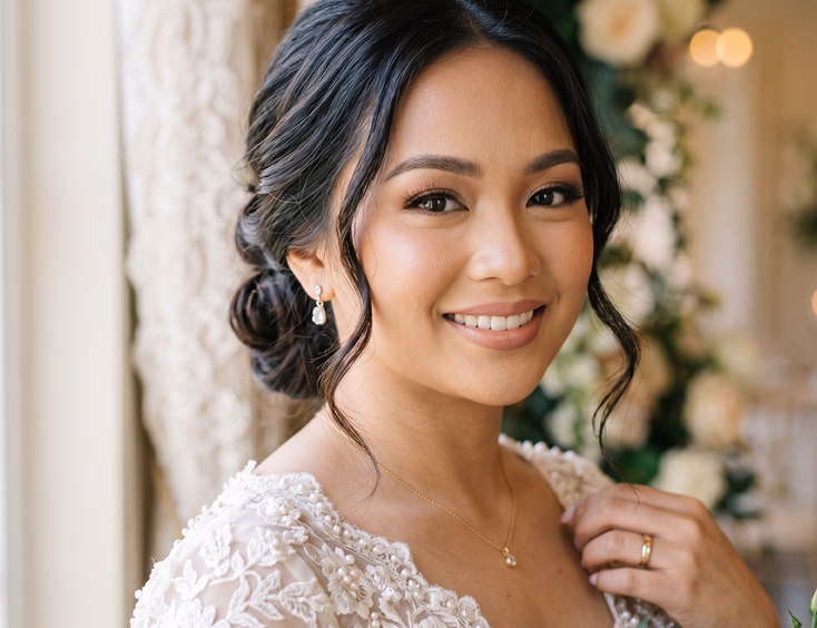 Close-up portrait of Filipino bride with soft glam makeup, warm neutral eyeshadow, wispy lashes, and nude pink lips with loose low bun in natural window light