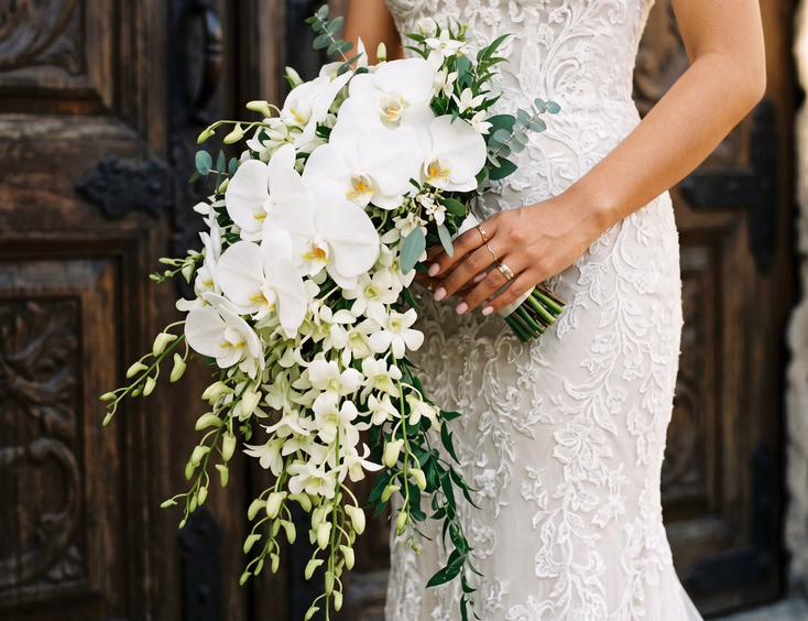 Close-up of a Filipino bride holding a cascading bridal bouquet of white Phalaenopsis orchids and trailing green Dendrobium orchid stems against a wooden church door in soft natural daylight