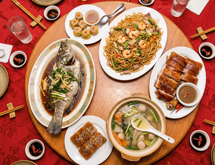 Overhead view of Filipino-Chinese tinghun feast spread on round table with lazy Susan featuring steamed lapu-lapu, pancit noodles, lechon, tikoy, and soup on red tablecloth