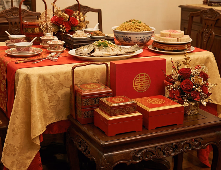 Tinghun side table in a warmly lit Filipino home sala displaying red and gold lacquered gift boxes with a floral arrangement, beside a formally set ceremonial table with traditional dishes including whole steamed fish, longevity noodles, and tikoy in red and gold table decor