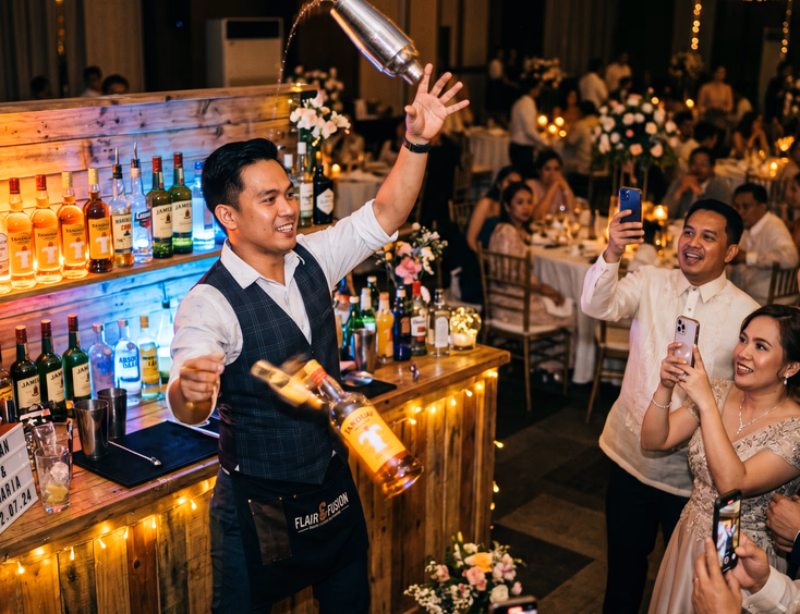 Filipino flair bartender tossing a cocktail shaker mid-performance at an evening wedding reception with guests watching and recording on their phones
