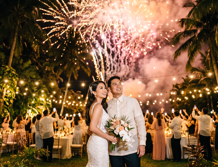 Filipino bride and groom in barong tagalog watching gold and pink fireworks at a tropical garden wedding reception.